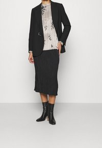 Donna che indossa un blazer nero, una blusa floreale chiara, una gonna midi plissettata nera e stivaletti neri con tacco, in piedi contro un muro bianco.