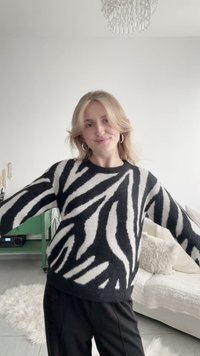 Joven con cabello rubio hasta los hombros, vistiendo un suéter de rayas de cebra en blanco y negro y pantalones negros, de pie en una sala de estar acogedora.