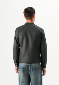 Jeune personne avec des cheveux bruns courts portant une veste noire et un jean bleu, se tenant de dos contre un fond blanc.