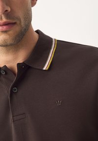 Brun polo med struktureret stof, der har en tre-knaps stolpelukning, kontrasterende stribe-detaljer på kraven og et lille emblem.