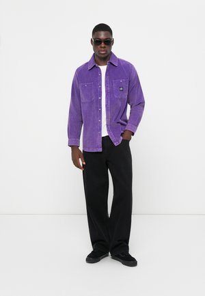 Purple corduroy shirt with buttons, two chest pockets, worn over a white t-shirt, paired with black wide-leg pants and black shoes.