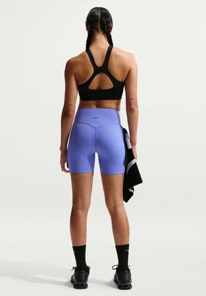 Frau mit geflochtenem Haar, die einen schwarzen Sport-BH und lila Nike-Shorts trägt, hält ein schwarz-weißes Handtuch und steht vor weißem Hintergrund.