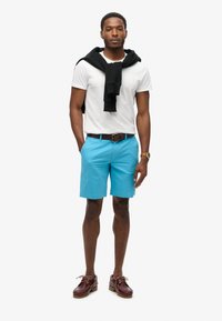 Camiseta blanca de manga corta, pantalones cortos turquesa, suéter negro caído sobre los hombros, cinturón marrón, zapatos de barco marrones con cordones, reloj dorado.