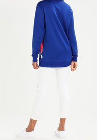 Blå sweatshirt med hög krage och sidodetaljer i rött och vitt, parat med vita leggings med slitna detaljer och ankellängd.