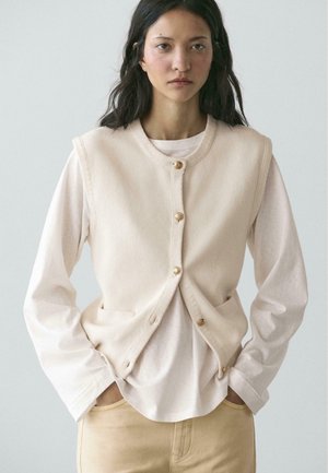 Jeune femme aux longs cheveux foncés portant un gilet beige boutonné par-dessus une chemise légère à manches longues et un pantalon beige, debout les mains dans les poches.