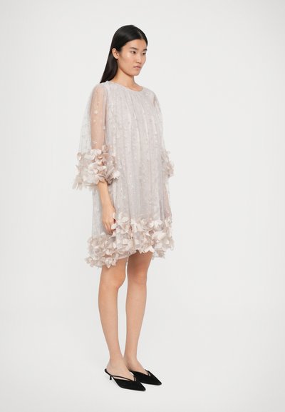 Robe florale rose pâle en tissu transparent, présentant une coupe ample, des appliqués floraux intricats sur les manches et l'ourlet, assortie de chaussures noires.