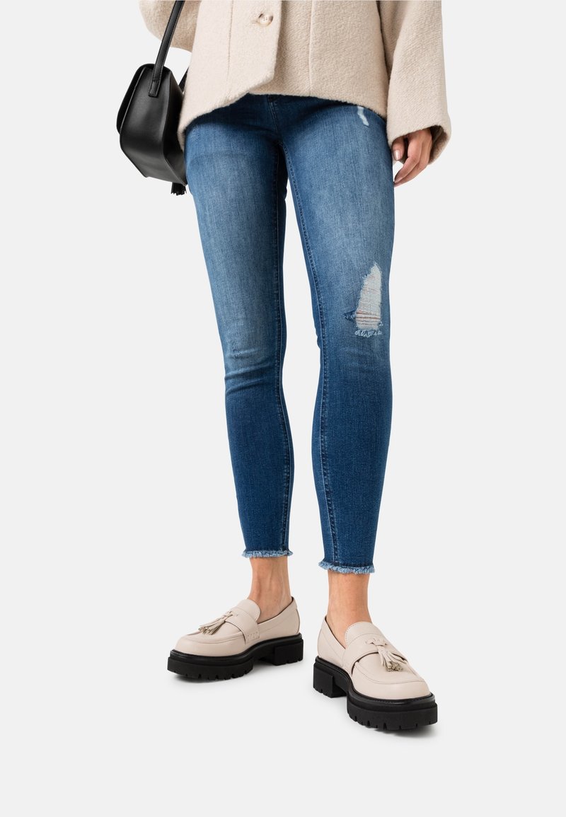 Personne portant des mocassins à pampilles beige, un jean skinny bleu déchiré et un manteau beige clair, tenant un sac à main noir à côté d'elle.