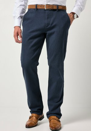 Mann trägt marineblaue Hose mit braunem Ledergürtel und passende braune elegante Schuhe, weißes Hemd und Uhr am linken Handgelenk.