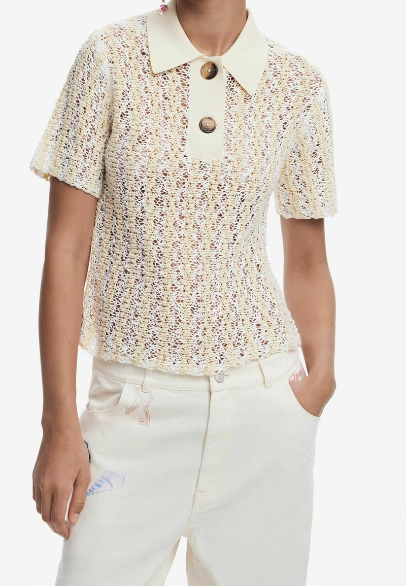 Femme portant un polo en crochet crème à manches courtes avec deux gros boutons, associé à un pantalon blanc avec une broderie discrète.
