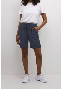 Pantaloni corti blu in cotone con fascia elastica in vita e lacci, dotati di due tasche e orli arrotolati, abbinati a una maglietta bianca e sneaker bianche.