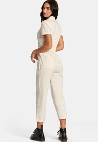Light beige jumpsuit with short sleeves and a fitted waist. It features a back pocket and cropped legs, paired with black lace-up boots.