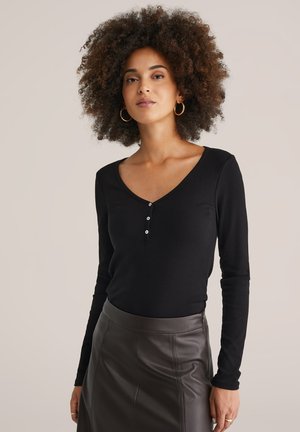Schwarzes Langarmoberteil mit V-Ausschnitt und drei Knopfdetails, kombiniert mit einem dunkelbraunen Lederrock. Glatte Textur, figurbetontes Design.