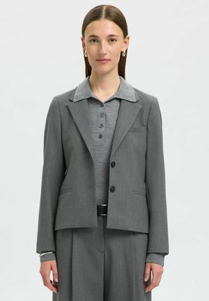 Blazer grigio su misura con revers, due bottoni e tasche laterali. Indossato sopra una camicia colletto grigio chiaro con patta dei bottoni.