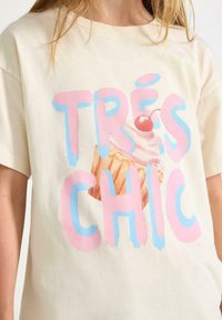 Gräddfärgad bomulls t-shirt med en cupcake-grafik med ett körsbär, tillsammans med texten "TRÈS CHIC" i stiliserad rosa och blå font.
