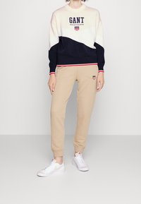 Pull tricoté à blocs de couleur marine et crème avec logo "GANT" ; pantalon de survêtement beige avec détail brodé ; baskets blanches.