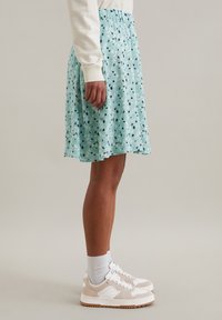 Jupe bleu clair, longueur genou, avec un motif floral et géométrique. Fabriquée en tissu doux avec une taille élastique pour le confort. Portée avec des baskets beiges.