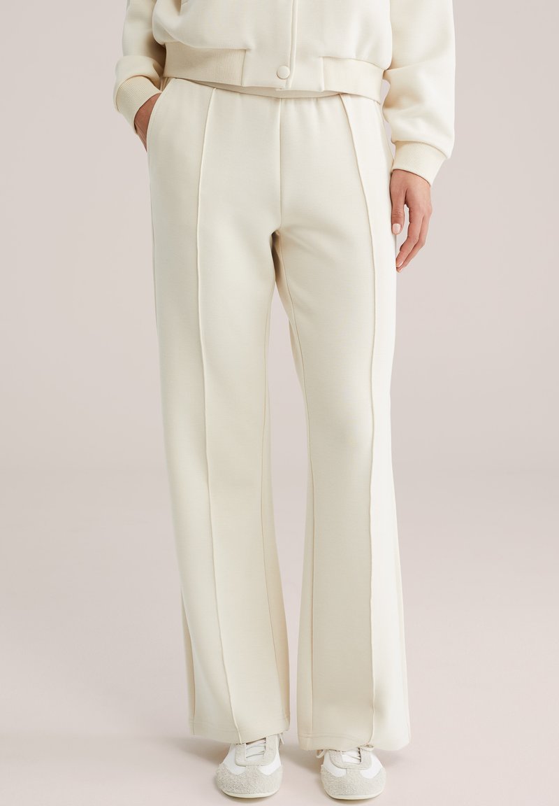 Bukser i cremefarve med vide ben og forreste sømme, båret sammen med matchende cremefarvet jakke og hvide sneakers, hænder delvist i lommerne.