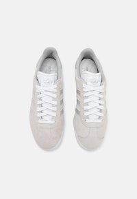 Adidași din suede gri deschis cu șireturi albe, având dungi argintii pe laterale și un guler căptușit. Talpă din cauciuc texturat.