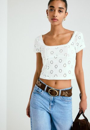 Jonge vrouw die een wit cropped top met oogkant draagt, lichtblauwe spijkerbroek met een bruine riem, en een bruine tas vasthoudt tegen een effen achtergrond.