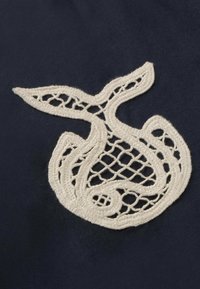 Motif en dentelle blanche brodée ressemblant à un poisson stylisé sur un tissu bleu foncé.