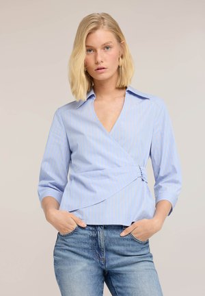 Blonde Frau trägt eine hellblaue gestreifte Wickelbluse mit Dreiviertelärmeln und blaue Jeans, Hände in den Taschen, neutraler Hintergrund.