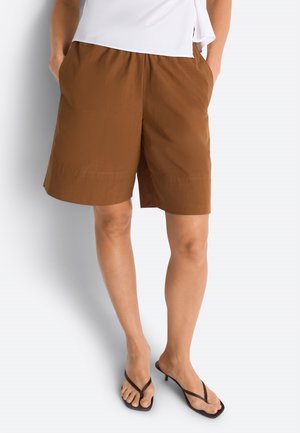 Kvinde iført løsthængende brune knælange shorts med sidelommer, hvid top og sorte sandaler med hæl, stående mod en lys baggrund.