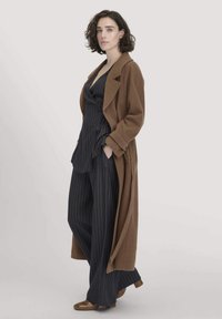 Cappotto lungo in lana marrone sopra una camicia navy a righe sottili e pantaloni larghi. Scarpe marroni, con una texture liscia e una vestibilità su misura.