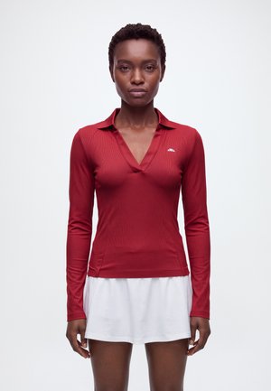 Une femme porte un haut rouge à manches longues et à col, ainsi qu’une jupe de tennis blanche, sur un fond clair et uni.