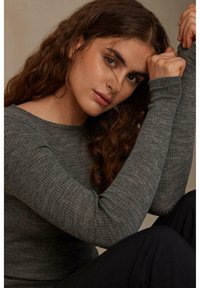 Šedý žebrovaný top s dlouhým rukávem a kulatým výstřihem. Modelka pózuje s překříženýma rukama, což ukazuje texturovanou látku a přiléhavý design. Měkké osvětlení.