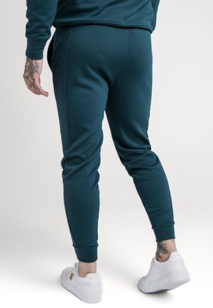 Man wearing teal jogger pants and white sneakers, standing with back facing the camera on a plain light background.