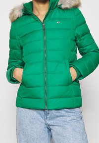 Chaqueta acolchada verde con cremallera frontal, bolsillos laterales y capucha forrada de pelo. Presenta costuras horizontales y puños de canalé.
