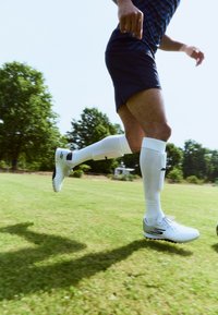 White athletic shoes with black accents, fitted black shorts, and white socks with protective shin guards, on grass, during motion.
