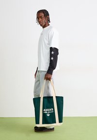 Junger Mann mit geflochtenen Haaren, der mehrlagige weiße und schwarze Ärmel mit Sternenmustern trägt, helle Jeans, schwarze Schuhe und eine grüne Adidas-Tote-Bag hält.