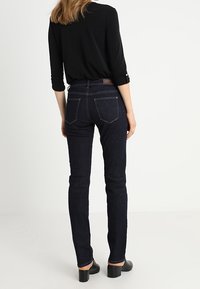 Jeans ajustados de mezclilla oscura con cintura alta, que presentan bolsillos traseros y detalles de costura sutiles. Combinados con una blusa negra y zapatos de tacón.