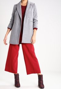 Cape grigio su misura con una texture liscia, abbinato a un maglione rosso e pantaloni larghi rossi. Stivali alla caviglia con motivo floreale completano l'outfit.