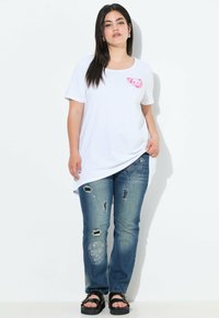 Camiseta blanca de algodón con un gráfico rosa, combinada con unos jeans azules desgastados que presentan bordados y rasgaduras, usados con sandalias de plataforma negras.