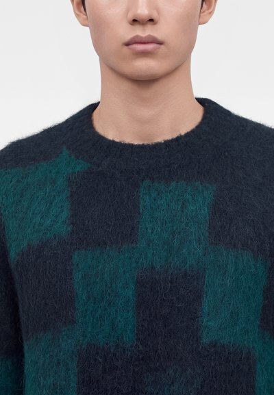 Gros plan d'une personne portant un pull bleu foncé duveteux avec de grandes formes géométriques bleu sarcelle, montrant le cou et le bas du visage sur un fond uni.