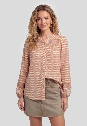 Vrouw met golvend haar, een blouse met patroon en knopen met bloemaccenten en een beige denim rok, glimlachend naar de camera.