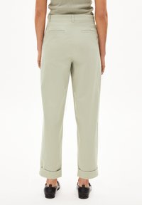Hellgrüne, hoch taillierte Hose mit umgeschlagenen Säumen und zwei Gesäßtaschen, aus einem glatten Stoff gefertigt. Sichtbare schwarze Slipper.