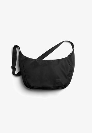 Sac bandoulière en nylon noir avec sangle ajustable et petite étiquette de marque rectangulaire sur le devant, sur fond blanc.