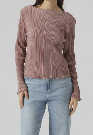 Femme portant un haut mauve à manches longues côtelé avec un col bateau et un jean en denim bleu clair, debout devant un fond uni.