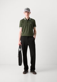 Grønn polo skjorte med hvit og svart stripet krage, svarte bukser, grå baseballcaps med logo, holder en svart bag.