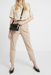 Camicia beige a maniche corte con bottoni e due tasche, abbinata a pantaloni tapered beige chiaro. Gli accessori includono una piccola borsa a tracolla in pelle nera lucida e ballerine nere.