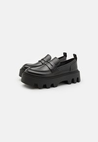 Mocassins en cuir noir à plateforme avec des semelles crantées épaisses et des tirettes au niveau du talon, présentés côte à côte sur un fond blanc.