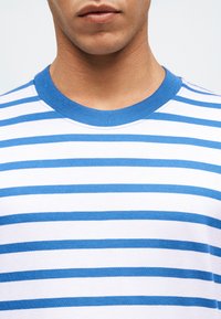 Mężczyzna w białej koszulce z poziomymi niebieskimi paskami i niebieskim prążkowanym okrągłym dekoltem, pokazany od szyi do klatki piersiowej.