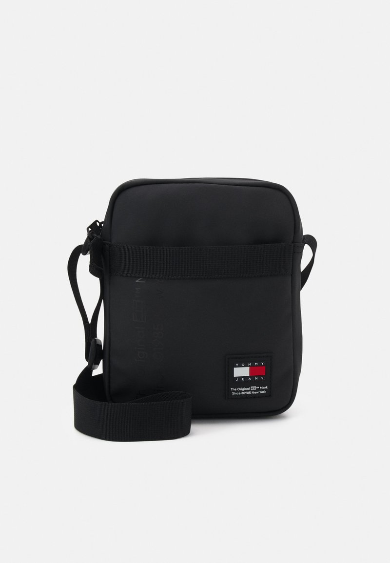 Borsa a tracolla nera realizzata in nylon morbido. Presenta una forma rettangolare, una tracolla regolabile e una toppa con logo sul fronte con accenti rossi, bianchi e blu.