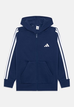 Námornícky zipsový mikina vyrobená z mäkkej tkaniny s bielymi pruhmi na rukávoch, dvoma prednými vreckami a kapucňou. Obsahuje logo Adidas v bielej farbe.