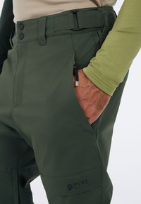 Groene broek met een soepele textuur, voorzien van twee knopen, zijzakken en een zaklogo. De riem heeft een zwarte hardware gesp.