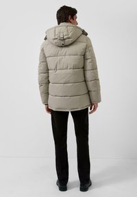 Šviesiai smėlinis pūkinis švarkas su kapišonu, kuriame yra siuvinėtas raštas ir sagtys. Pora su juodomis kelnėmis ir tamsiais batais, matoma iš nugaros.