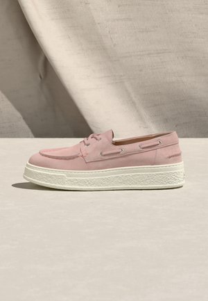 Roze suède bootschoen met witte, gestructureerde rubberen zool, veters en stiksels op een neutrale beige ondergrond en stoffen achtergrond.